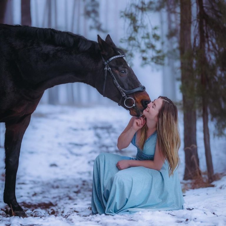 Eine Frau in einem blauen Kleid sitzt im Schnee und küsst ein herabschauendes Pferd.