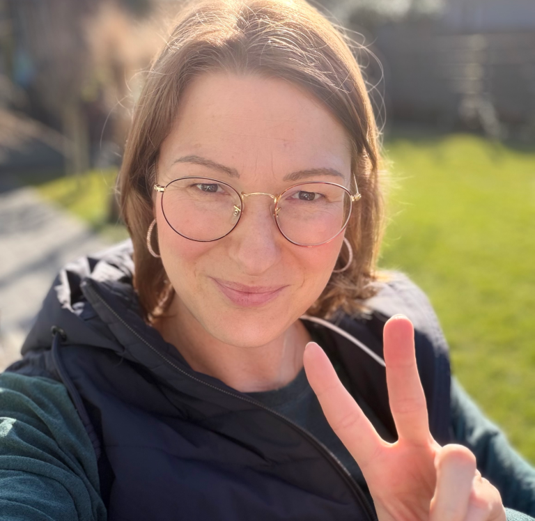 Kathi Stec mit Brille und kurzer Haarfrisur zeigt ein Peace-Zeichen im Freien.