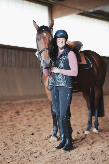 Kathi Stec in einem Stall steht neben Lusitanostute. Sie trägt einen Helm und Reitkleidung.