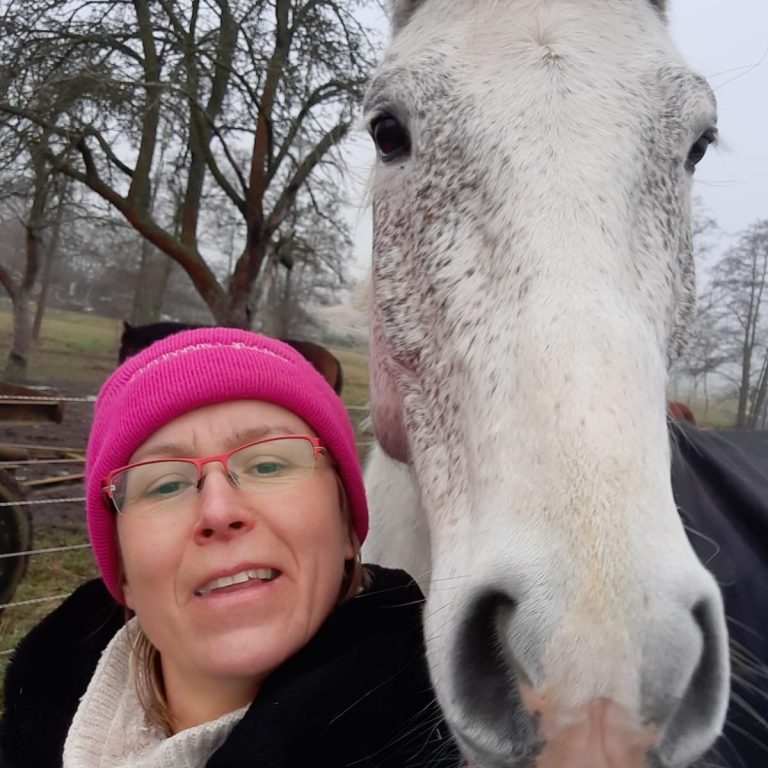 Frau mit rosa Mütze posiert zusammen mit einem weißen Pferd.