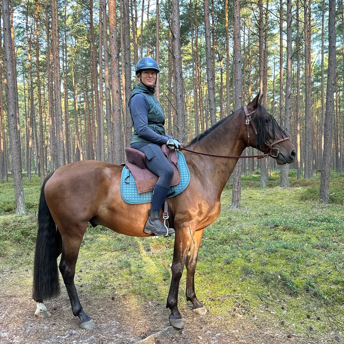 Kathi Stec auf ihrem braunem Pferd im Wald, umgeben von hohen Bäumen.