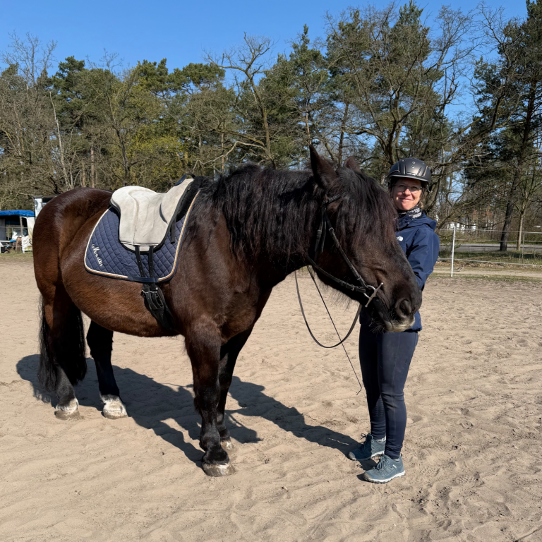 Reiterin mit einem schwarzen Voriger auf einem Sandplatz, umgeben von Bäumen.